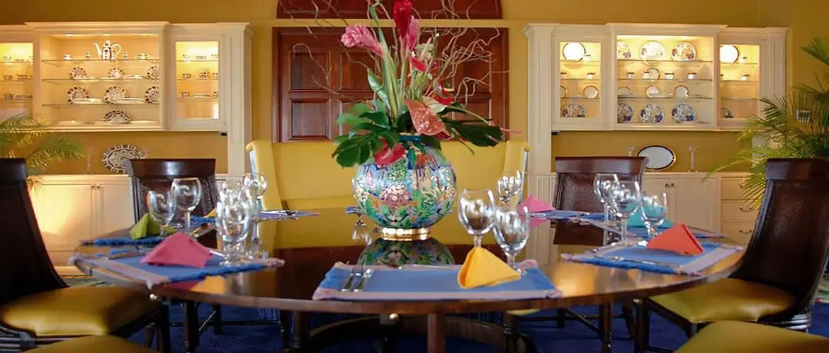 Yellow chairs and a woven banquette surround a dark wood table set with colorful napkins, where a tall vase of tropical flowers centers the room between illuminated china cabinets.