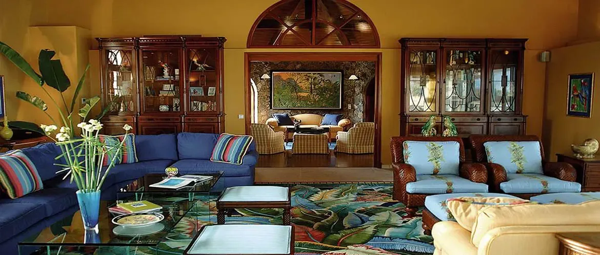 A navy L-shaped sofa with striped pillows sits on a bold tropical-leaf rug, where wicker armchairs and glass cabinets flank an arched doorway with a sunburst carving.