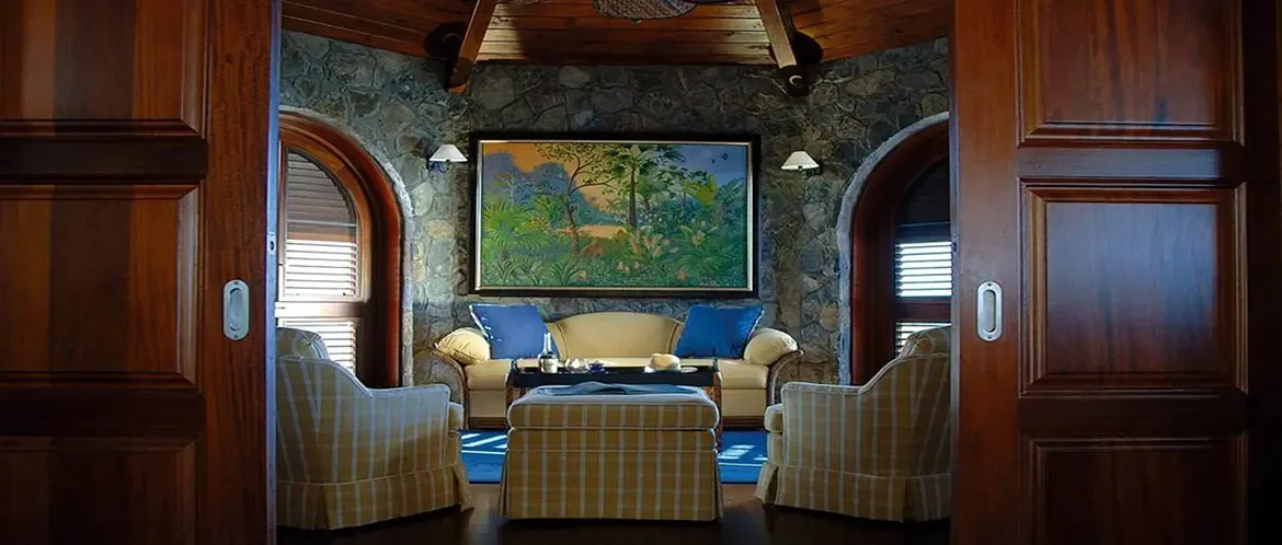 Yellow checkered armchairs and a cream sofa sit in a stone-walled octagonal room under a vaulted wood ceiling, where a tropical landscape painting hangs between arched shuttered windows.