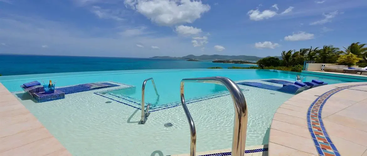 Chrome handrails enter a turquoise infinity pool with blue mosaic lounging platforms, where a champagne bottle and two glasses sit beside an ocean that merges seamlessly with the horizon.