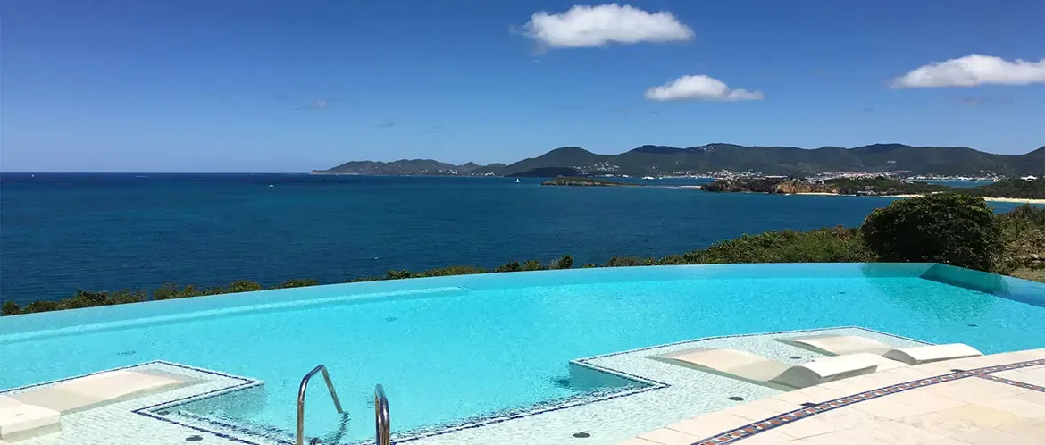 White loungers sit submerged on a turquoise pool sun shelf with blue mosaic edges, where the infinity rim merges with a sapphire ocean and distant green hills along the coast.