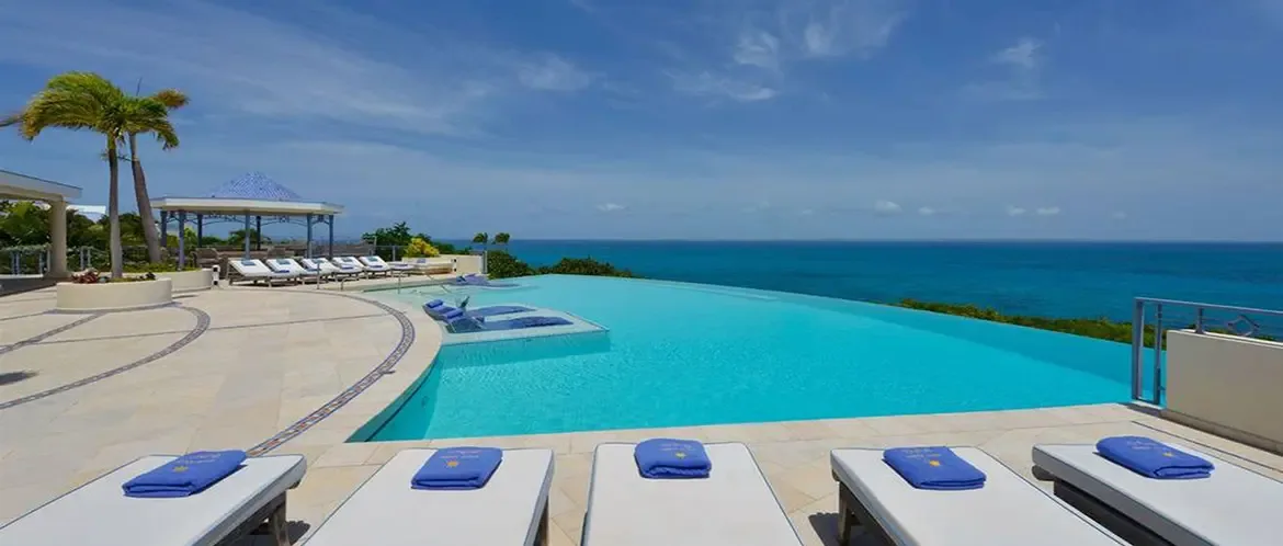 Two submerged blue loungers sit on a turquoise pool sun shelf beside chrome handrails, where the infinity edge merges with a deep blue ocean and distant green islands.