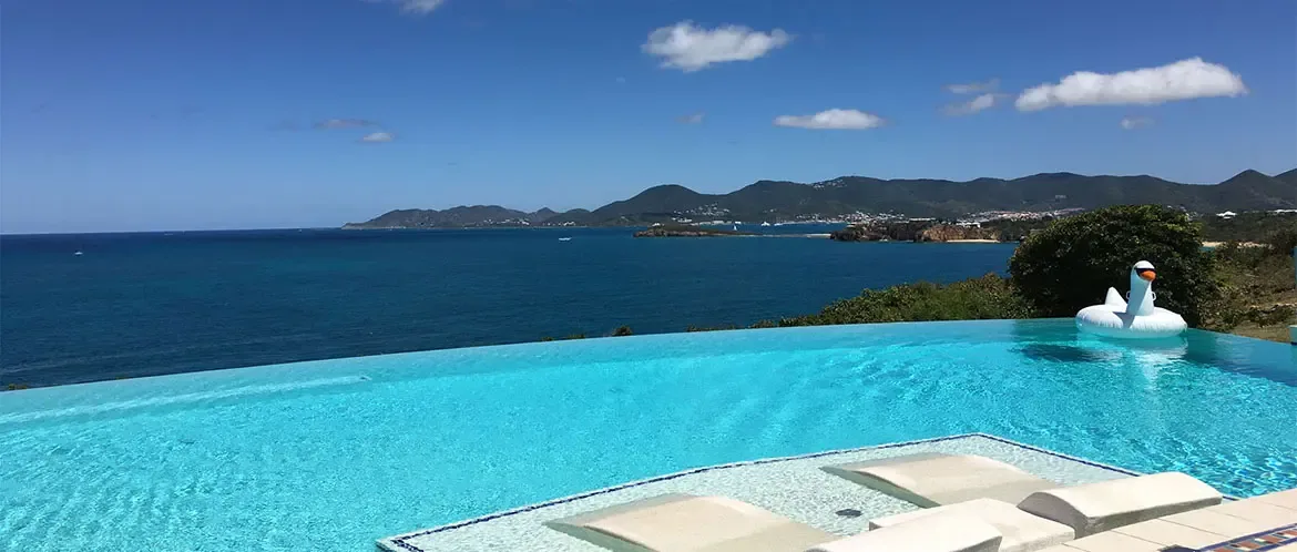Three white loungers sit submerged on a turquoise pool sun shelf beside a swan inflatable, where the infinity edge meets a deep blue ocean stretching to distant green islands.