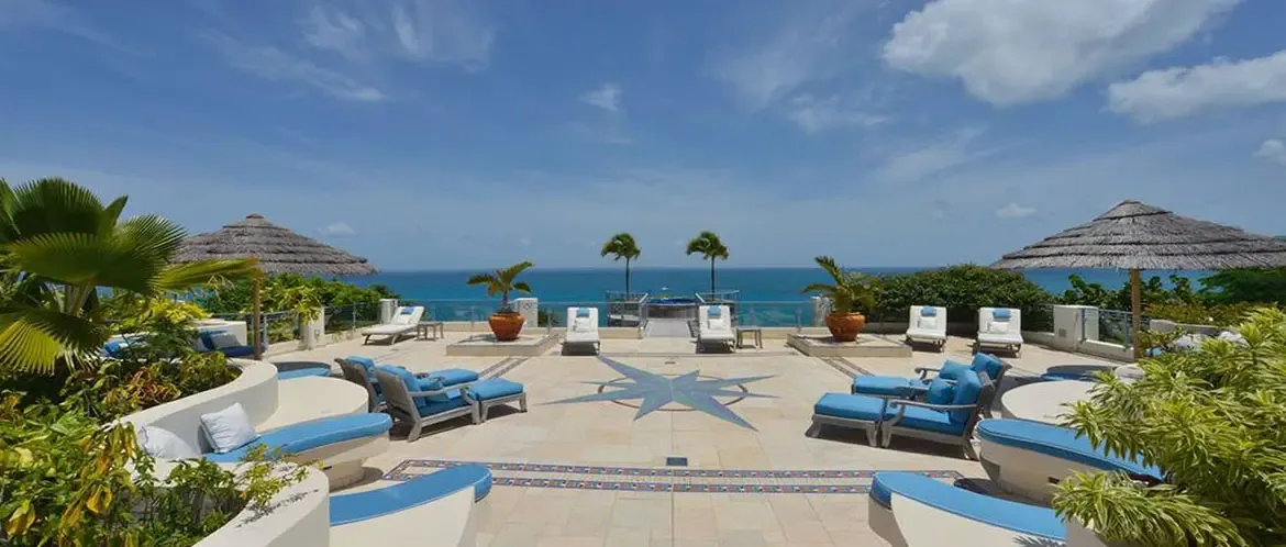 Blue-cushioned curved benches sit on a stone terrace with a compass rose mosaic, where thatched palapas shade wooden armchairs beside a glass railing and turquoise ocean.