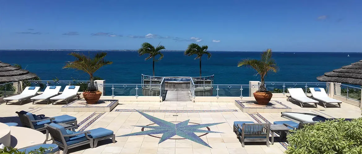 Blue-cushioned wooden chairs sit on a stone terrace with a compass rose mosaic, where palapas and white loungers frame a circular hot tub overlooking a turquoise ocean.