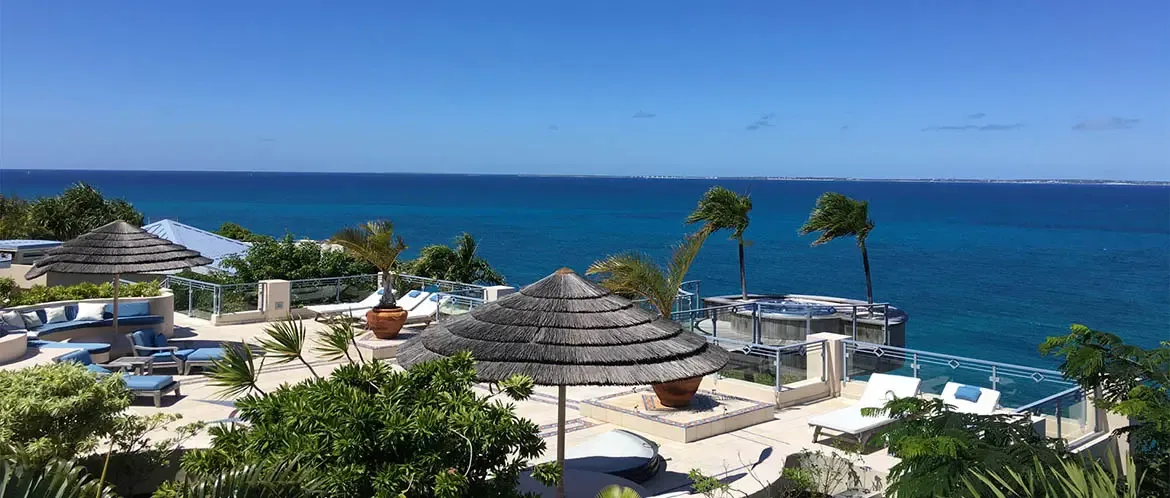 Blue-cushioned curved benches and wooden chairs sit under thatched palapas on a stone terrace, where a circular hot tub overlooks a turquoise ocean stretching to distant islands.