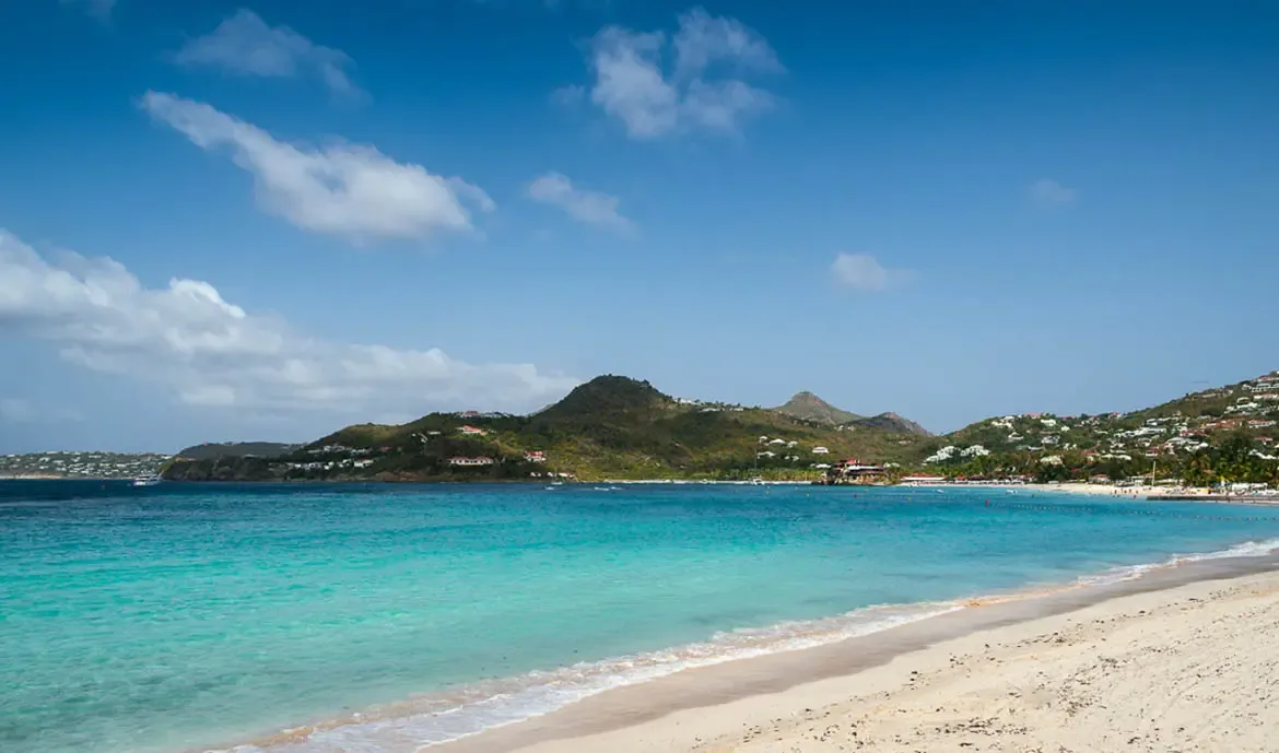 An aerial view captures a pristine white sand beach and turquoise water, where red-roofed villas nestle between lush green hills and a Caribbean bay.