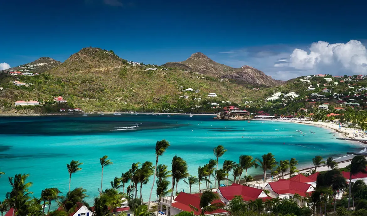 An aerial view captures red-roofed villas nestled among palms above turquoise Saint Jean Bay, where Eden Rock perches on a rocky outcrop and green hills rise across the water.