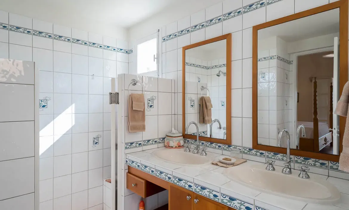 A white-tiled double vanity with oval sinks and wood-framed mirrors reflects the bedroom beyond, where blue-and-white decorative tile borders and shell-motif accents create a coastal bathroom.