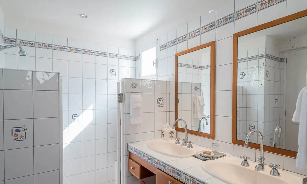 A white-tiled double vanity with oval sinks and chrome faucets sits under wood-framed mirrors, where blue-and-white decorative tile borders and face-motif accent tiles add Mediterranean charm.