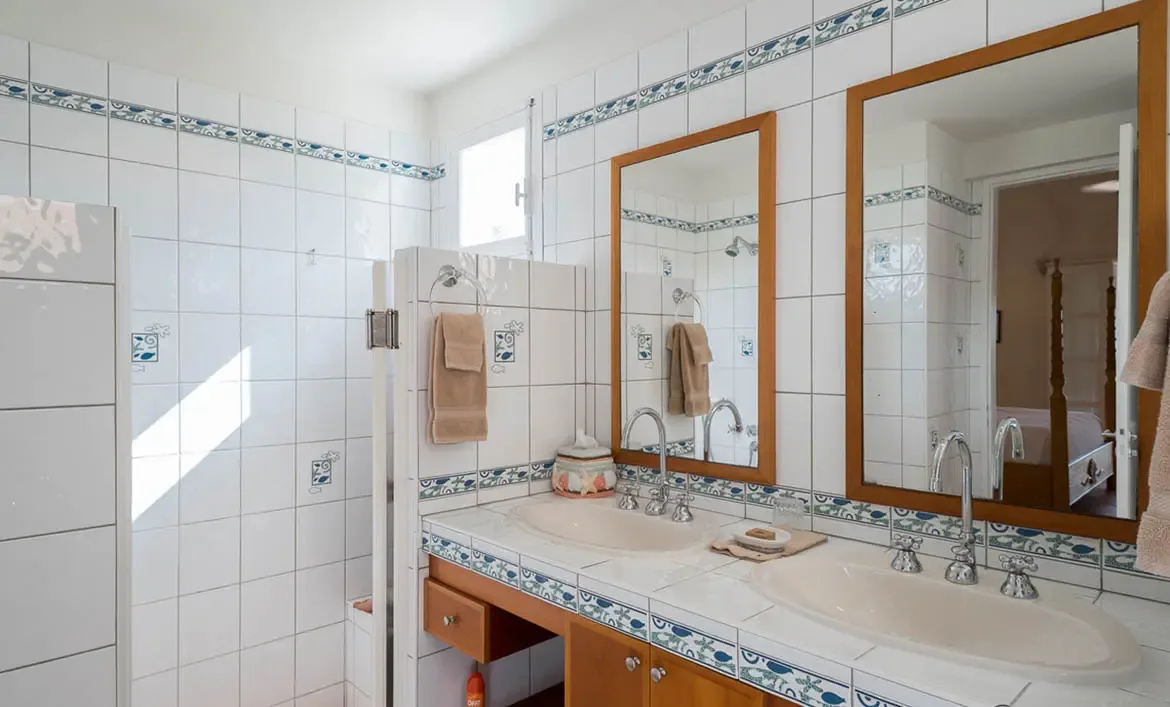 A white-tiled double vanity with goose-neck faucets and wood-framed mirrors sits beside a tiled shower partition, where blue decorative tiles and a coral jar add seaside character.