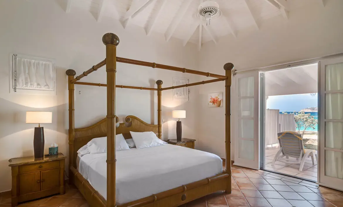 A bamboo four-poster bed with white linens sits on terracotta tiles under a vaulted ceiling, where open French doors lead to a patio with a lounge chair overlooking turquoise ocean.