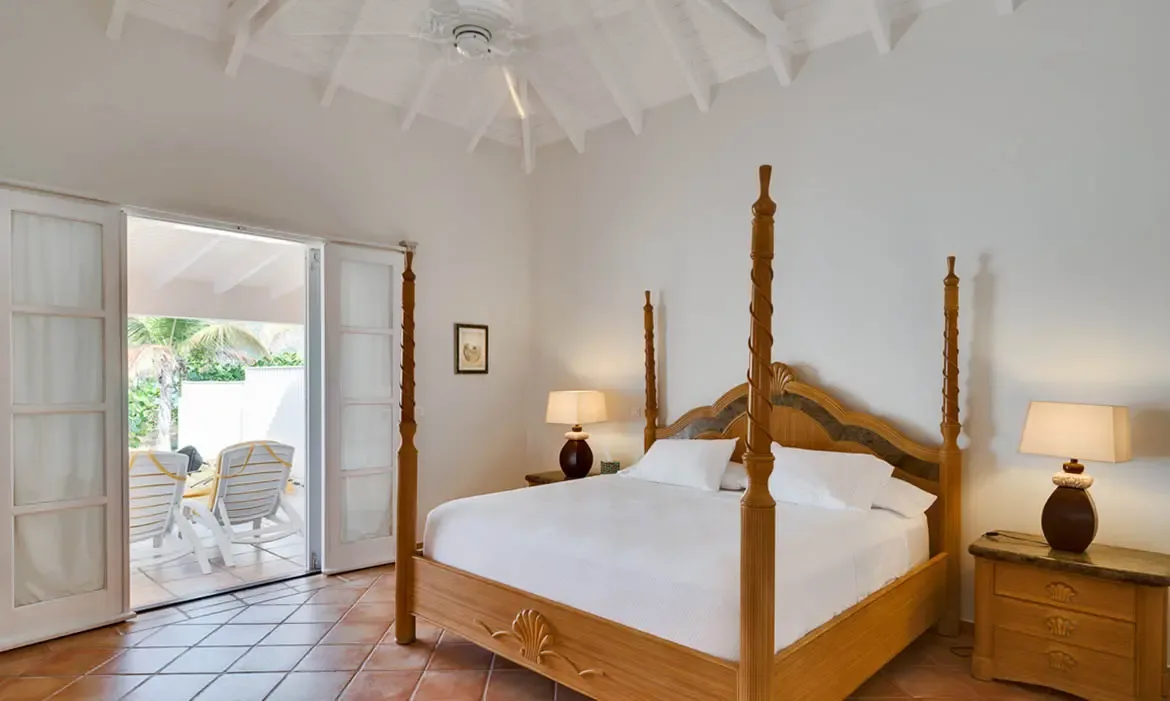 A carved wood four-poster bed with shell-motif headboard and white linens sits on terracotta tiles, where open French doors reveal palm trees and a sun-drenched patio.