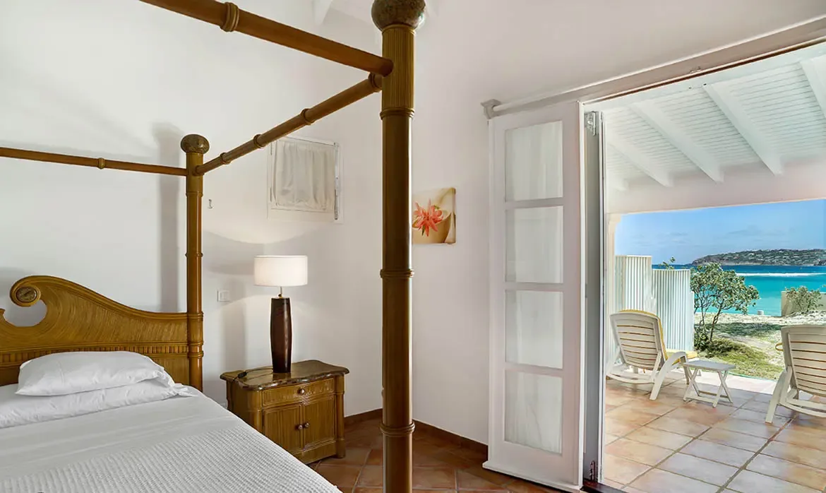 A bamboo four-poster bed with white linens sits before open French doors, where a balcony with loungers overlooks turquoise ocean and green hills.