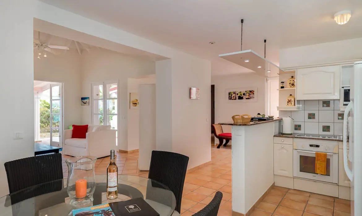 A round glass table with wicker chairs and a rose bottle sits beside a granite breakfast bar, where white cabinets and an archway to a living room with red pillows complete the open layout.