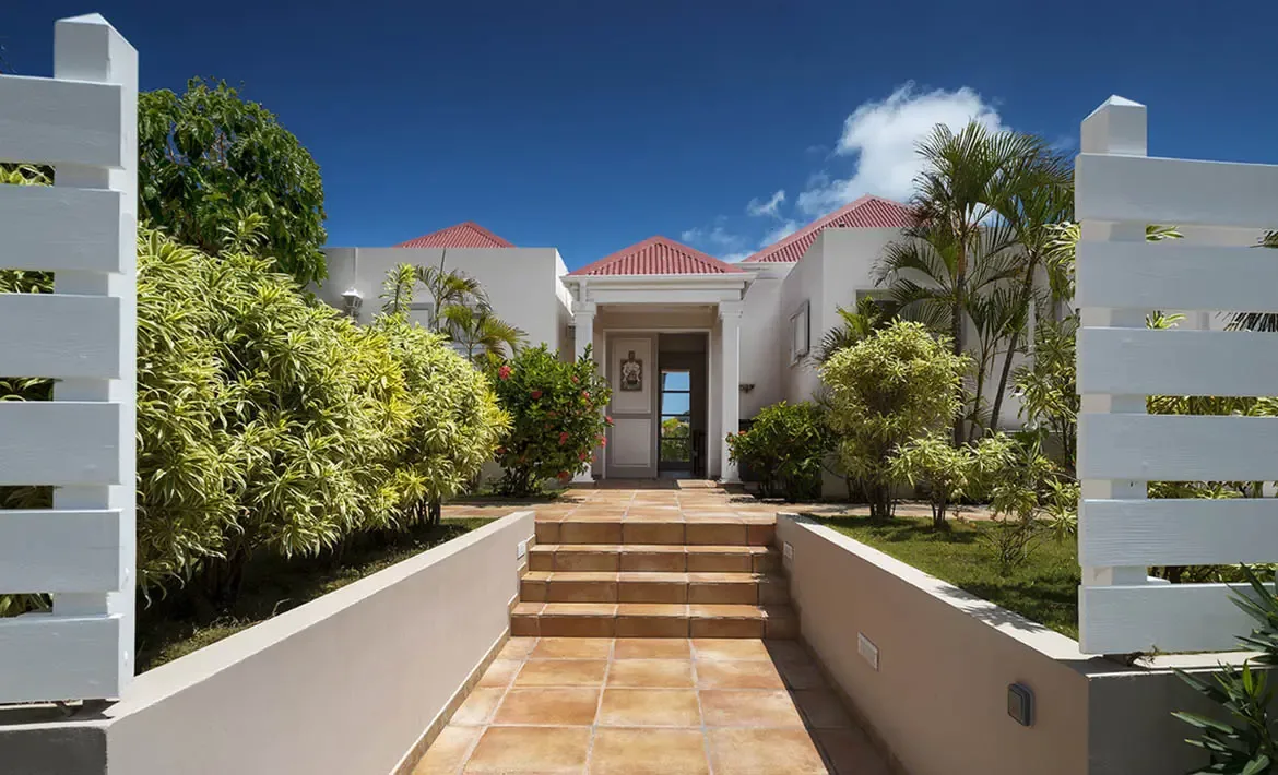 White columns frame a wide terracotta path leading into a red-roofed villa, where lush yellow bushes and palm trees flank the entrance under a deep blue sky.