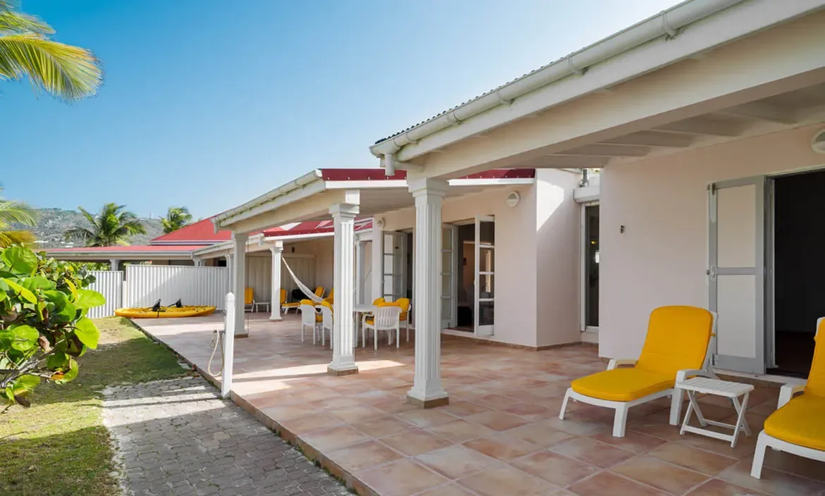 Yellow loungers and a dining terrace sit under a deep veranda with white columns, where a yellow kayak rests on a lawn and palms reach toward a blue sky.