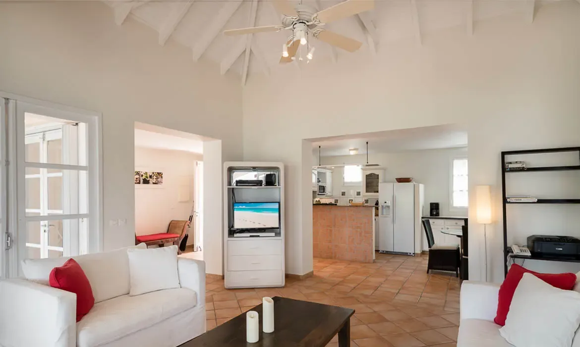 White sofas with red pillows and a TV facing a candlelit coffee table sit under a vaulted ceiling, where a kitchen with white cabinets connects to the open living space.