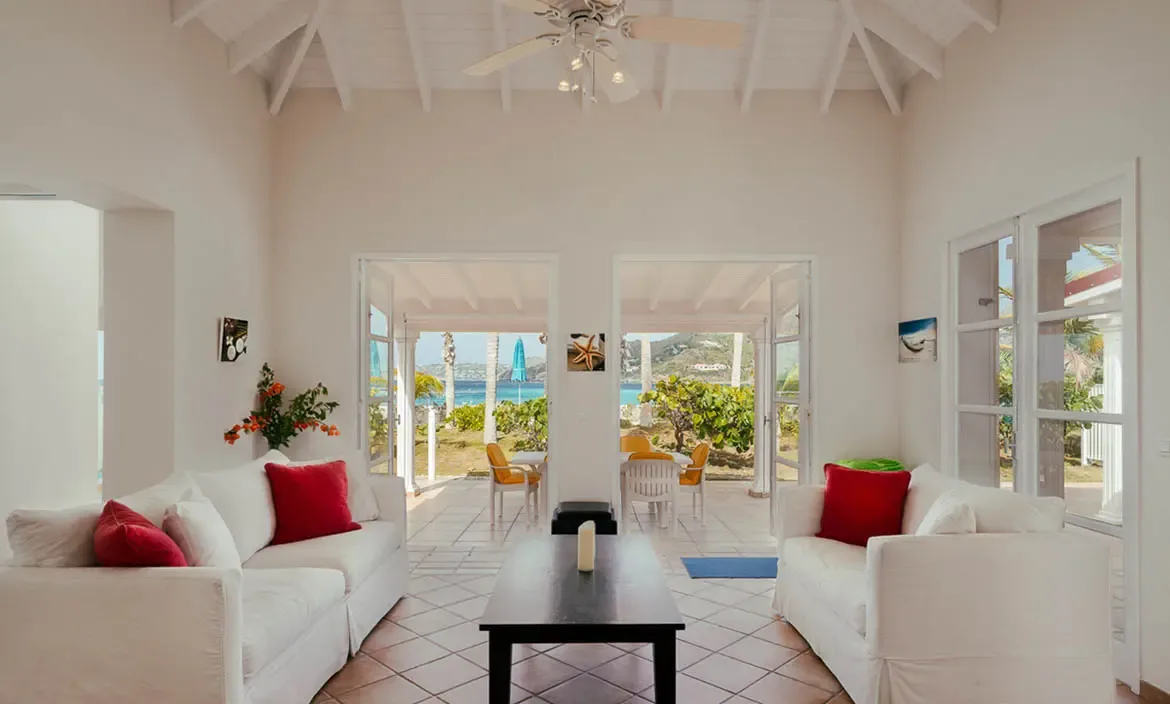 Two white sofas with red pillows sit on terracotta tiles under a vaulted ceiling, where open French doors reveal a yellow dining set and turquoise bay beyond.