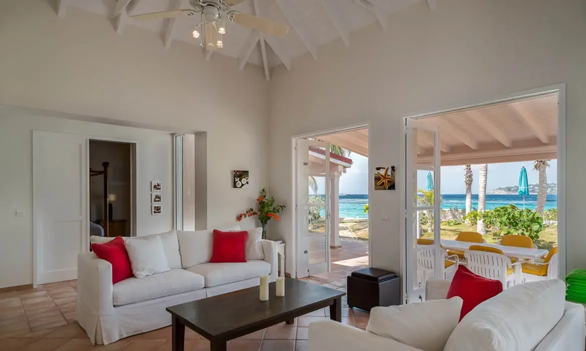 White sofas with red pillows sit under a vaulted ceiling on terracotta tiles, where open French doors lead to a yellow-cushioned dining terrace and turquoise ocean.