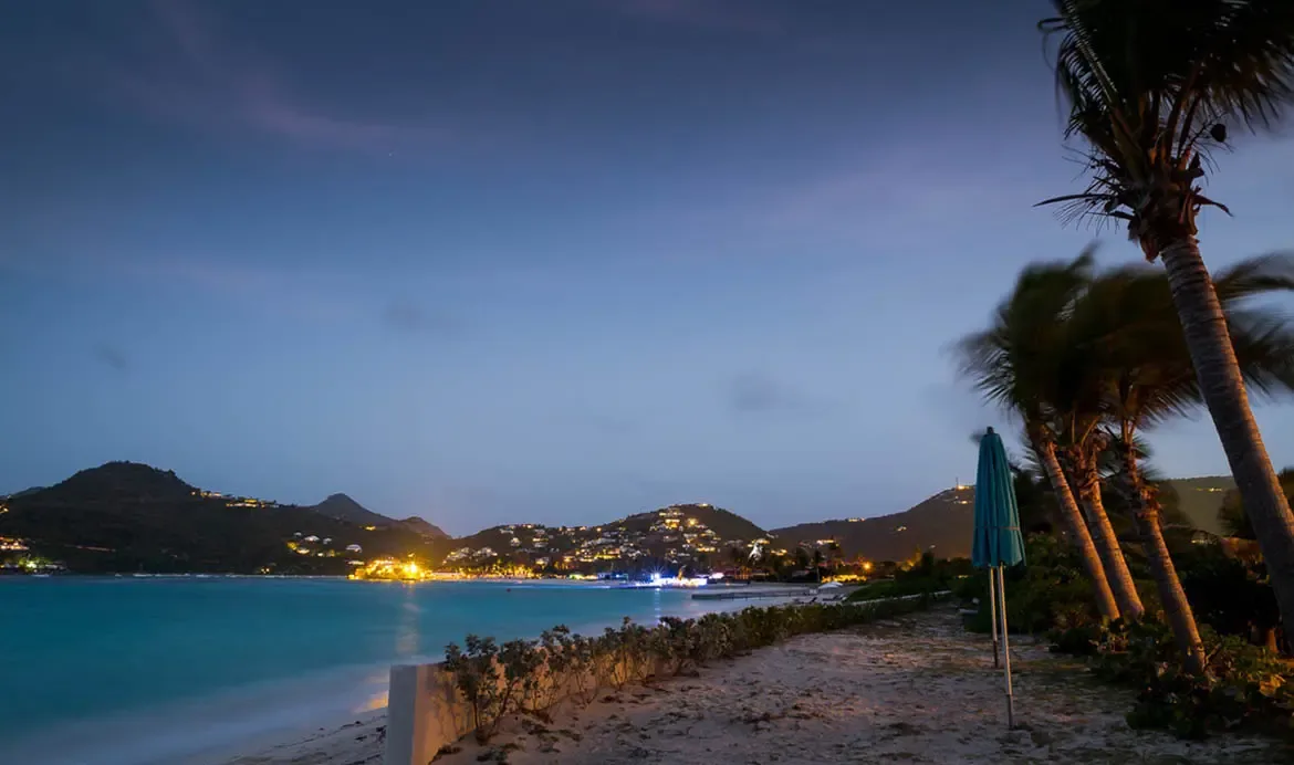 At twilight, swaying palms and a closed teal umbrella frame a sandy path, where a calm turquoise bay reflects distant hillside lights under a deep indigo sky.