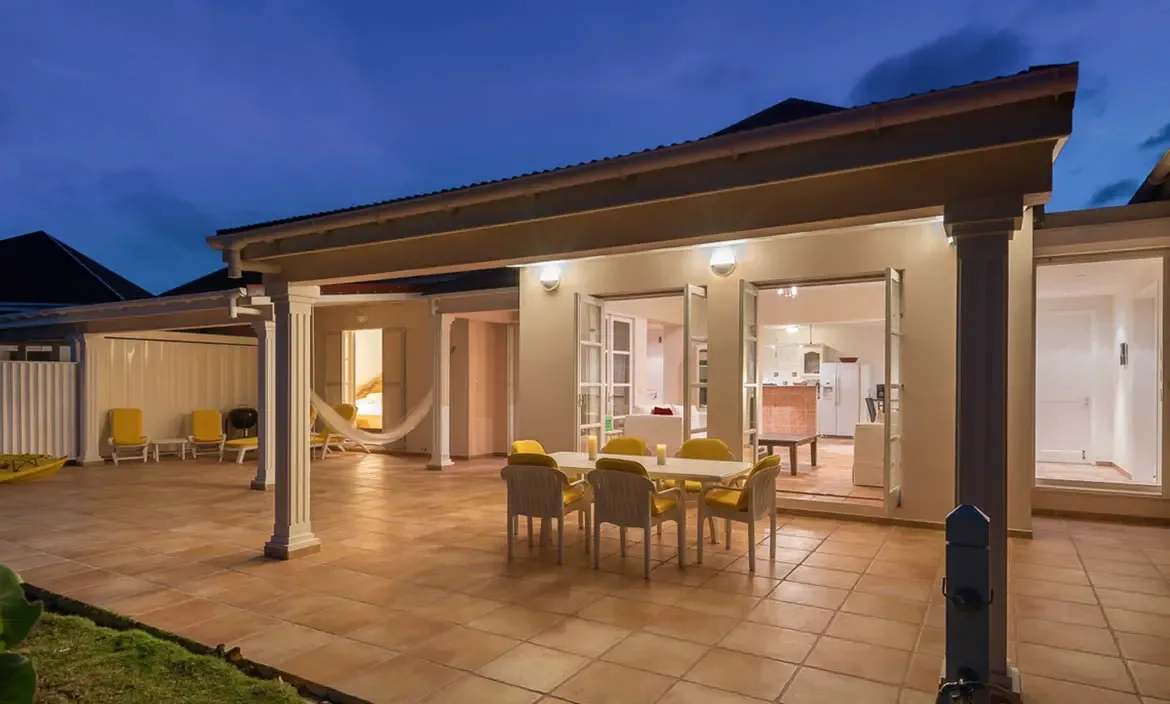 Yellow-cushioned dining chairs surround a white table on a lit terrace, where a hammock swings between columns and a white kitchen glows through open French doors.