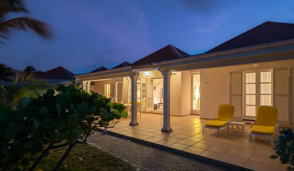 At blue hour, white columns and a deep porch warm golden beside red pyramidal roofs, where interior light spills onto a terracotta terrace under an indigo sky.