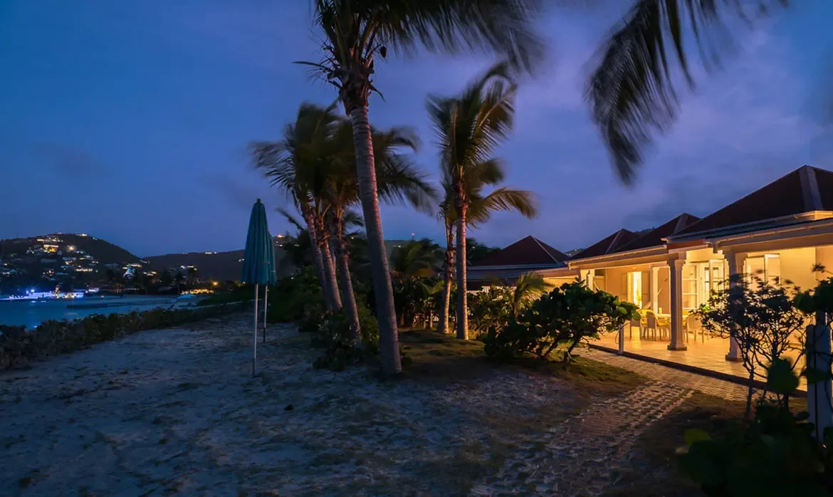 At blue hour, a white villa glows warmly beside swaying palms, where a turquoise beach umbrella and a distant hillside of lights meet a calm bay.