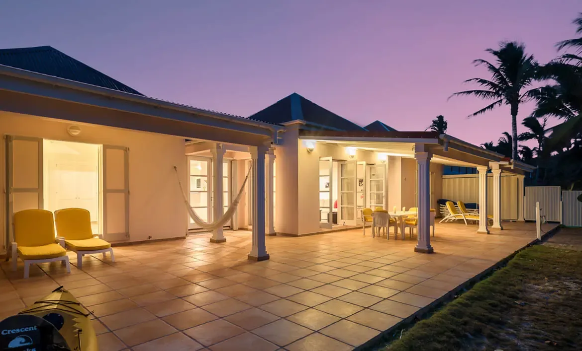 At twilight, a white villa glows warmly through open French doors, where yellow loungers and a dining terrace sit under a deep porch framed by palm silhouettes.