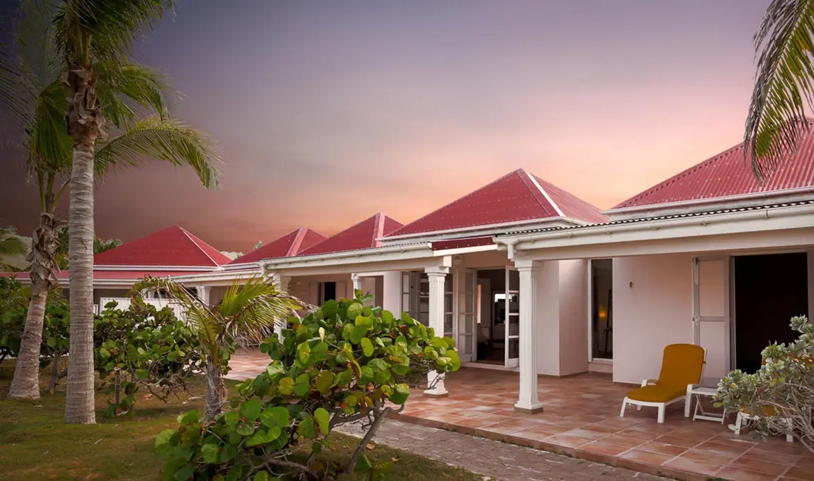 A bright yellow lounger sits on a terracotta patio beside a white villa with red pyramidal roofs, where palm trees and sea grape frame a twilight ocean view.