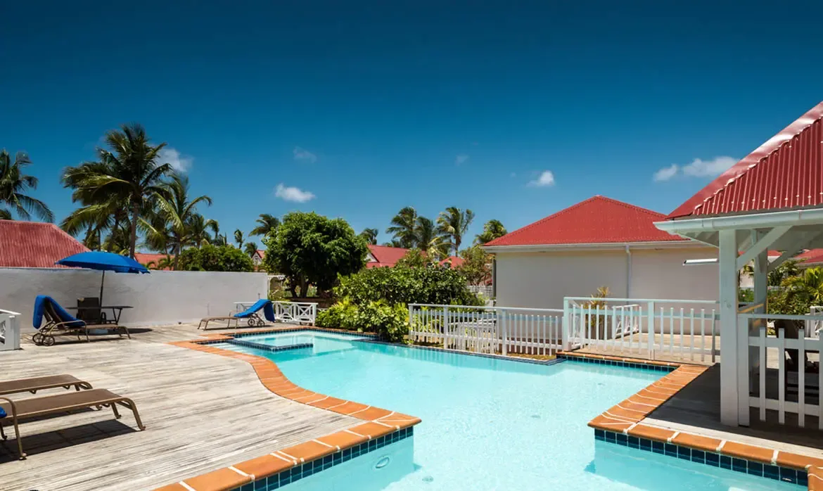 A turquoise freeform pool with blue tile edges sits on a weathered deck beside a white villa with red corrugated roofs, where a blue umbrella shades loungers under palm trees.