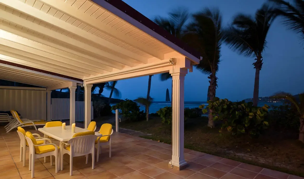 A white dining table with yellow-cushioned chairs sits under a slatted pergola with fluted columns, where golden lights and palm trees frame a twilight ocean view.