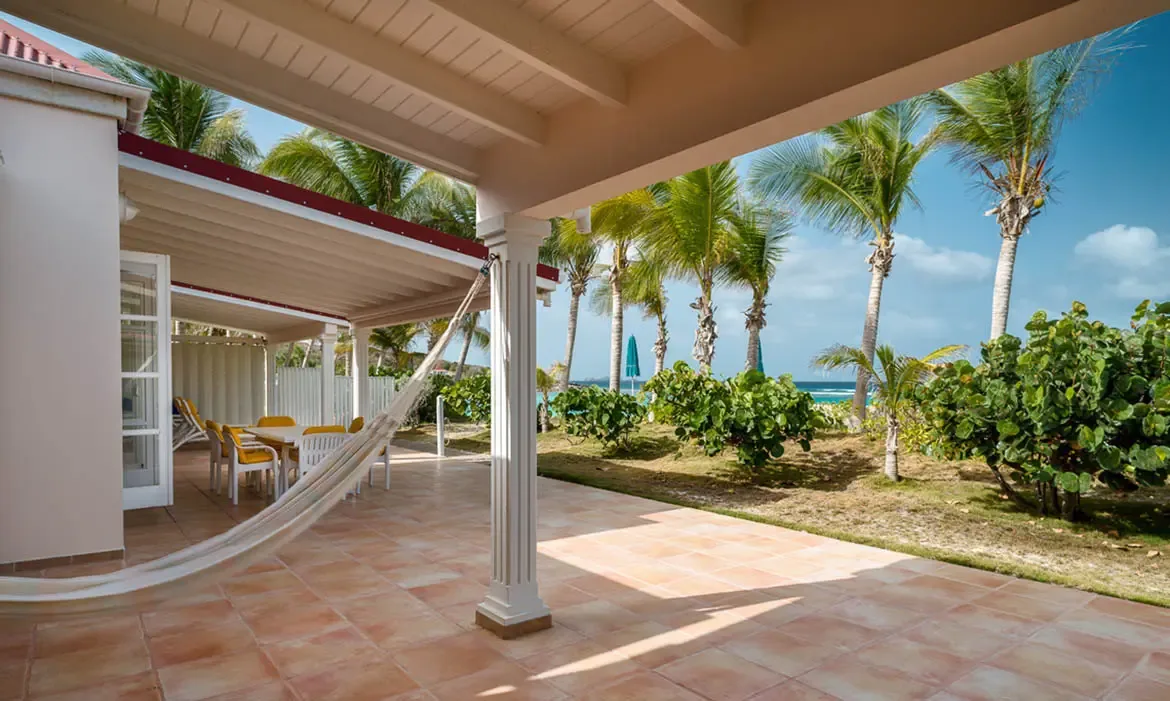 A white hammock stretches between columns on a terracotta terrace, where yellow-cushioned dining chairs and lush palms overlook turquoise ocean under a clear sky.