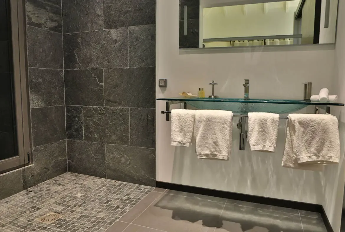 A glass vessel sink floats on a sleek glass shelf with chrome fixtures and neatly hung white towels. Dark stone tiles with natural veining line the shower area while a large rectangular mirror reflects the clean minimalist design—contemporary luxury in every detail.