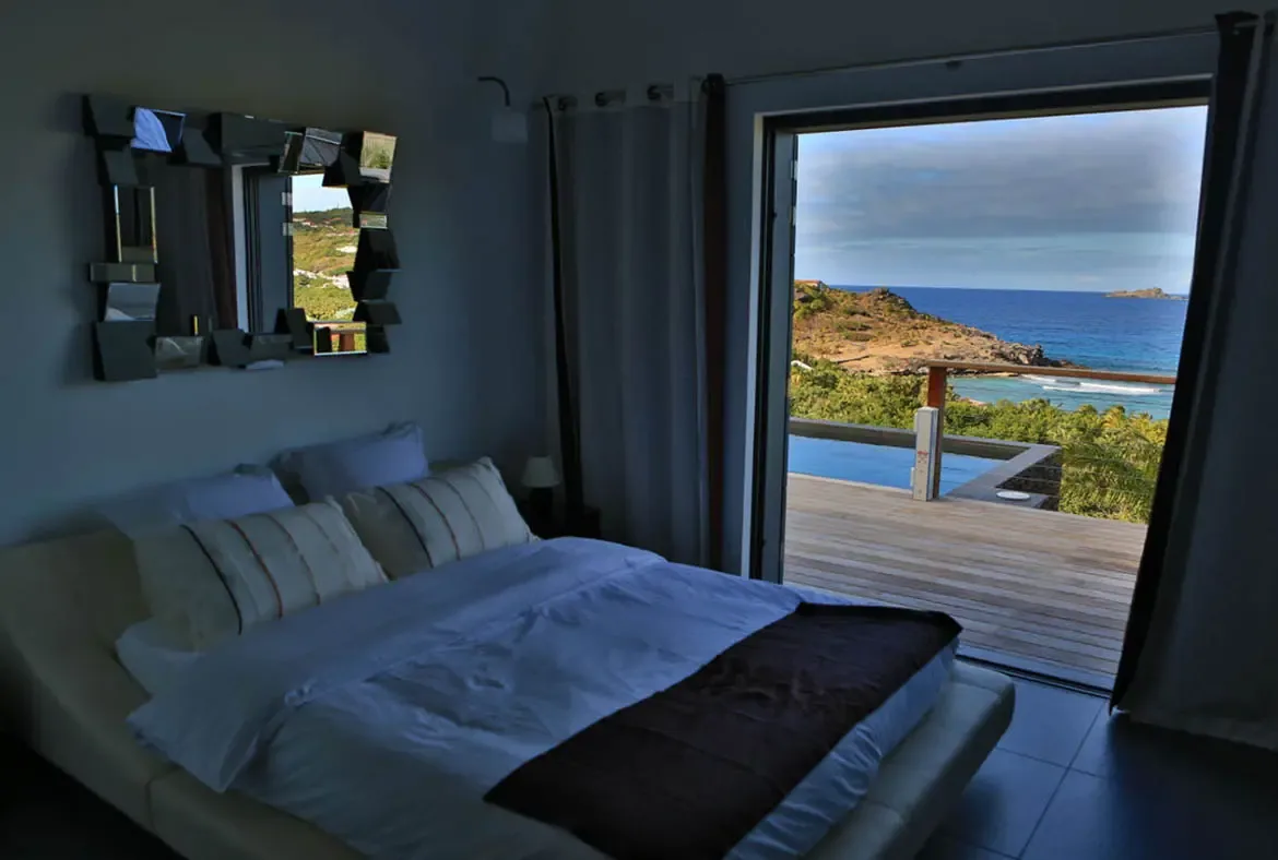 A white-dressed bed with striped accent pillows faces a breathtaking ocean panorama visible through wide doors. A geometric mirror of small rectangles adds artistic flair to the gray wall while the wooden deck leads to views of rocky cliffs and distant islands—serenity meets drama.