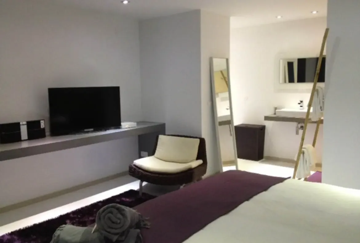 A white-dressed bed with a purple throw sits above a plush purple shag rug, while a wicker armchair with cream cushions faces a sleek console and flat-screen TV. Beyond the open partition, a floating sink and bamboo towel rack reveal the modern bathroom—contemporary comfort in every corner.