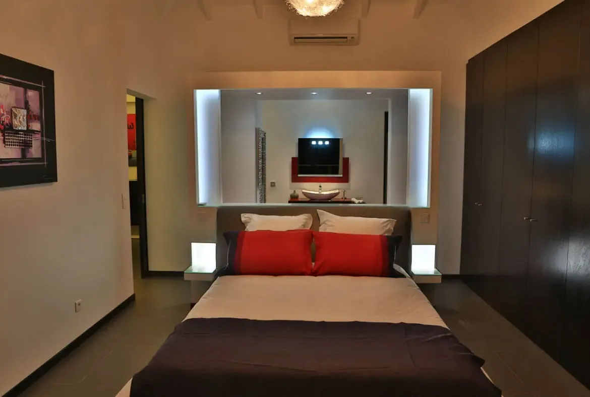Bold red accent pillows pop against crisp white linens on a bed flanked by glowing white nightstands with integrated light panels. An illuminated mirrored wall behind reveals a vessel sink and a red mirror, while a textured chandelier adds warmth—bold, modern, unforgettable.