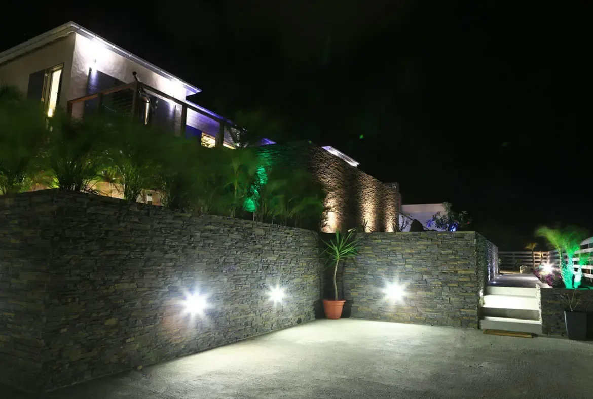 Stacked stone walls and lush landscaping glow under green uplights and warm wall sconces at this modern villa. Illuminated white stairs lead to the upper level while terracotta potted plants add organic texture—nighttime elegance that makes arriving feel like an event.