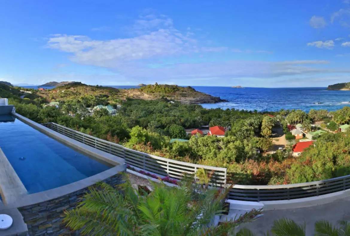 A sleek infinity pool with crystal blue water perches above lush green hills dotted with red-roofed villas. Beyond the tropical canopy, the deep blue ocean meets rugged peninsulas under a bright Caribbean sky—this is St. Barths from above.