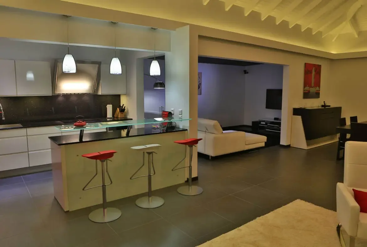 A sleek open-concept kitchen with white cabinets and a black island features red and white bar stools that echo the bold red artwork on the far wall. Beyond, a light sectional faces a wall-mounted TV against lavender walls—modern Caribbean living with a pop of color.