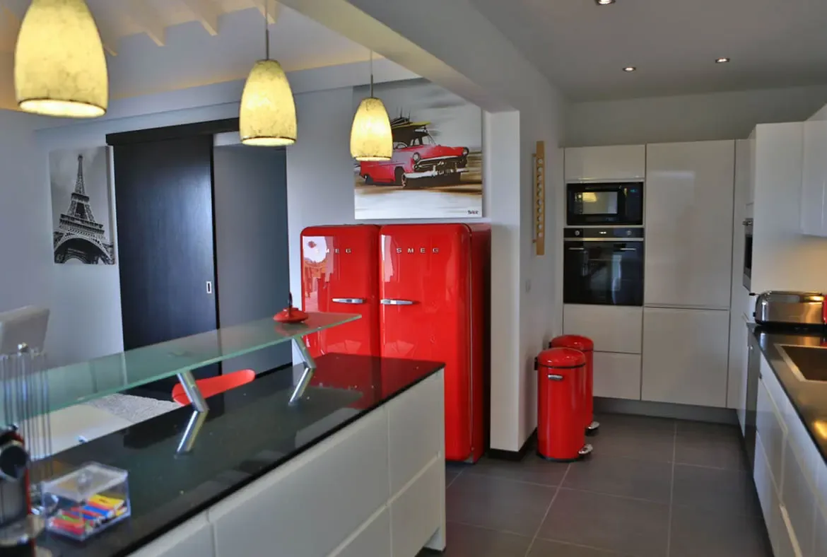 A vibrant red SMEG refrigerator and matching red trash cans pop against sleek white high-gloss cabinets and a black island countertop. A black-and-white Eiffel Tower print and the red car artwork tie the retro-meets-modern theme together—this kitchen has personality.