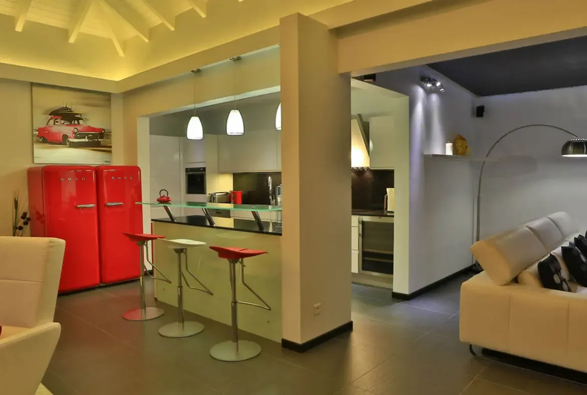 Two vibrant red SMEG refrigerators steal the show in this sleek kitchen, echoed by a framed red car artwork above. Red and white bar stools line the glass-topped island while pendant lights cast a warm glow—bold Caribbean style meets modern functionality.