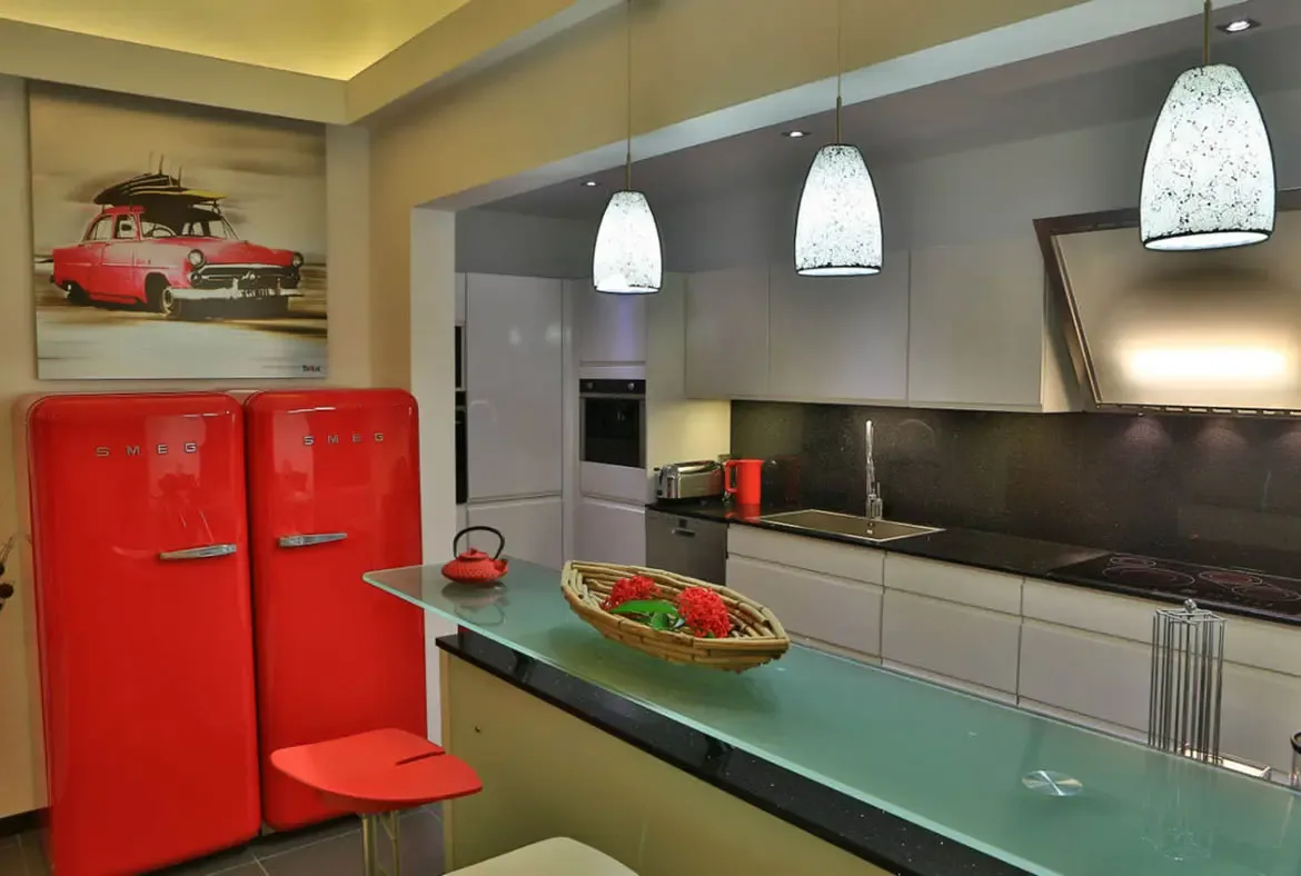 Two iconic red SMEG refrigerators anchor this vibrant kitchen, echoed by a wall print of a red classic car with surfboards on the roof. A translucent green glass island countertop, speckled pendant lights, and a wicker basket of red flowers complete this retro-meets-modern Caribbean kitchen.