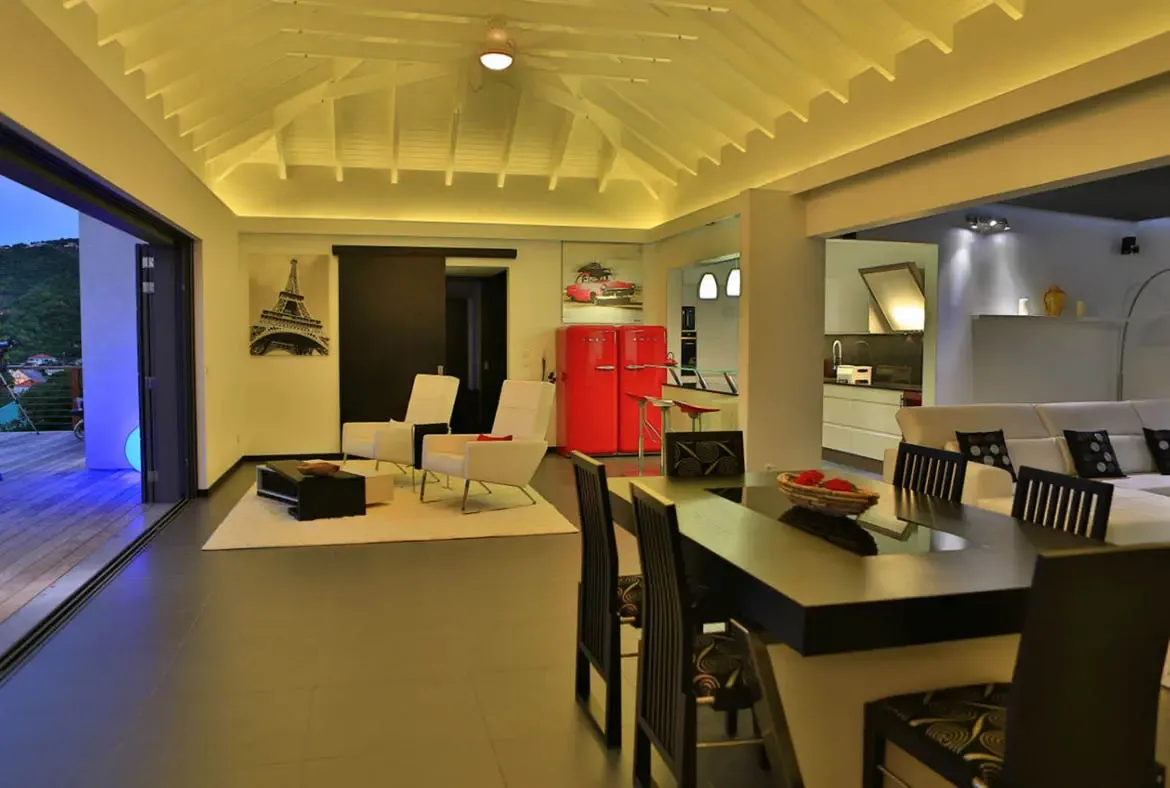 Warm recessed lighting glows across this open-concept space where white armchairs, a dark dining table with red fruit, and the iconic red SMEG fridge come together. Beyond the glass doors, lush green hills fade into dusk—day transitions to evening in this stylish Caribbean retreat.