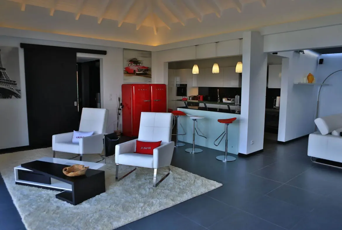 White armchairs—one with a red "St Barth" cushion—sit on a plush rug before a geometric coffee table. Beyond, the red SMEG fridge and matching bar stools anchor the kitchen beneath white pendant lights—this villa wears its Caribbean identity proudly.