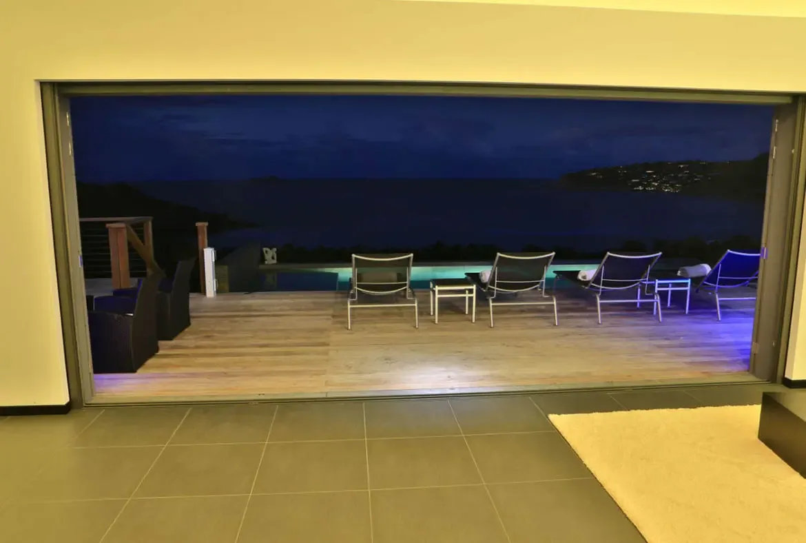 The open-concept living room flows straight onto a wooden terrace where lounge chairs and wicker seating face the dark ocean. Blue and purple LED lights trace the deck edge while distant coastal lights twinkle across the water—indoor-outdoor living under the Caribbean stars.