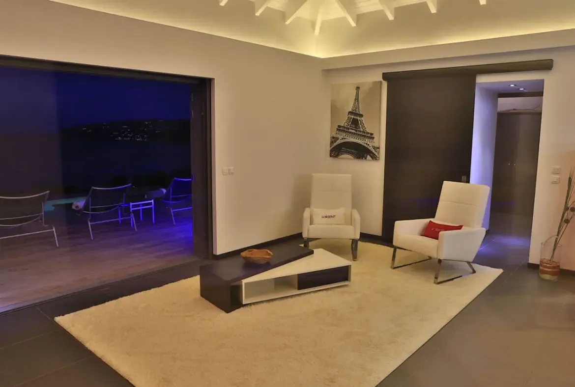 Two white armchairs with red and graphic pillows sit on a light rug before a striking black-and-white Eiffel Tower artwork. The open-concept space reveals a terrace glowing with blue LED lights and distant city lights across the water—Parisian chic meets Caribbean nights.