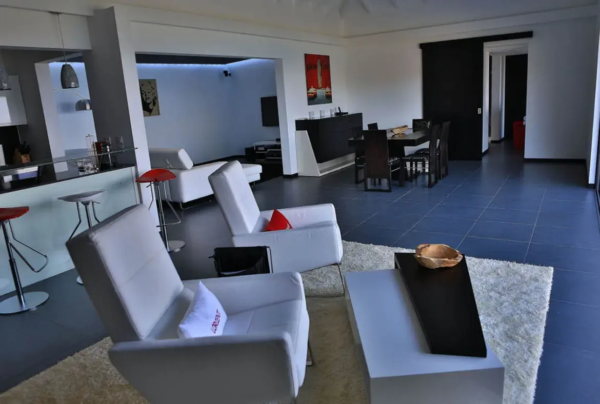 White leather armchairs with red accent cushions sit on a shaggy rug before a geometric coffee table. Red bar stools line the glass-topped kitchen counter while a pop-art portrait and red cityscape artwork add bold personality—minimalist design with maximum Caribbean flair.