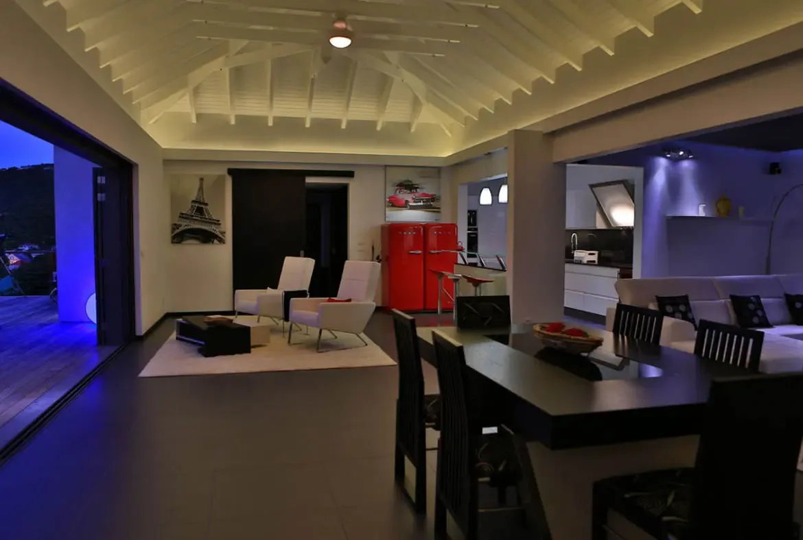 The open-concept living space comes alive under vaulted white-beamed ceilings: white armchairs, a dark dining table with a bowl of red fruit, and the unmistakable red SMEG fridge unite the zones. Beyond the glass, blue terrace lighting hints at evening cocktails awaiting on the deck.