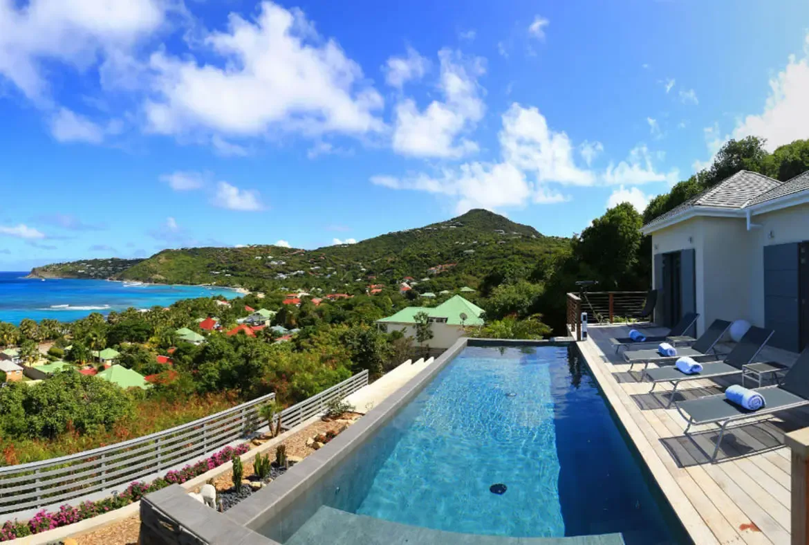 A sleek rectangular infinity pool with crystal blue water sits on a wooden deck with lounge chairs and rolled blue towels. Beyond, rolling green hills dotted with colorful homes cascade toward a turquoise shoreline—your private perch above the Caribbean with views that never end.